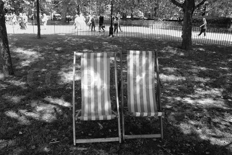 Two deck chairs under the tree and people are walking in Hyde Park in the city London in England on a sunny day in spring in black and white, stock photo