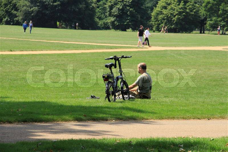 Walking people and a man is sitting on the lawn with the bike nearest him in Hyde Park in London in England on a sunny day in spring, stock photo