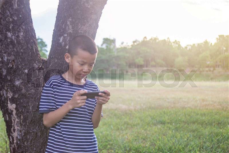 Young cute asian boy using a smartphone to play game in the park without care the surrounding, stock photo