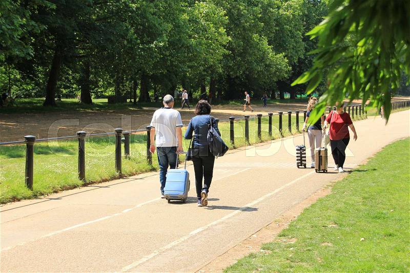 Couple, man and wife with trolly suitcase and an oncoming couple are walking at Hyde Park in London in England on a sunny day, stock photo