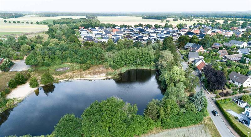 Aerial view of a suburb behind a small lake and a wooded area, with detached houses, semi-detached houses and terraced houses with small front gardens and green lawns in northern Germany, stock photo