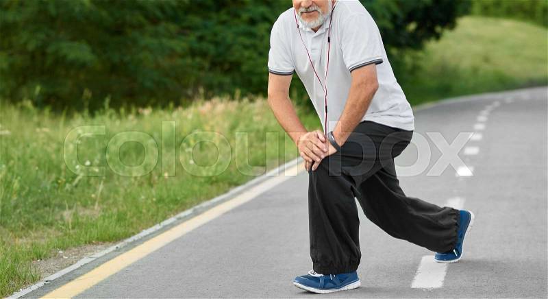 Cropped photo of training senior man wearing white beard, white polo shirt, black trousers and blue sneakers. Doing exrcises on race track in city park. Healthy lifestyle, fit, sporty figure, stock photo