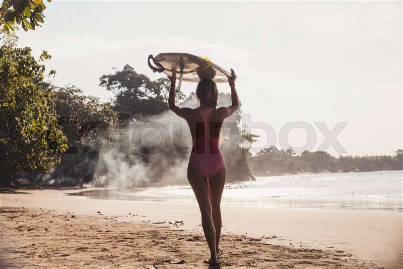 Back view of sportswoman carrying surfing board on head while walking on coastline, stock photo