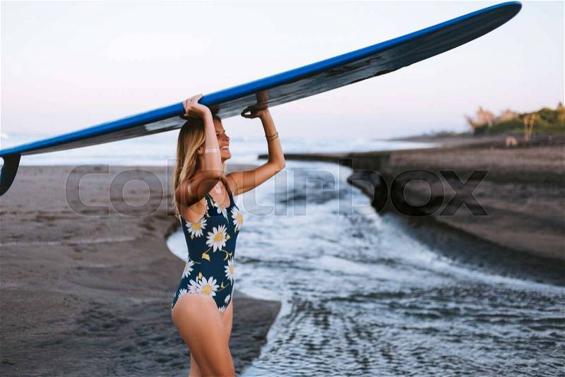 Young smiling sportswoman in swimming suit carrying blue surfing board on head on coastline, stock photo