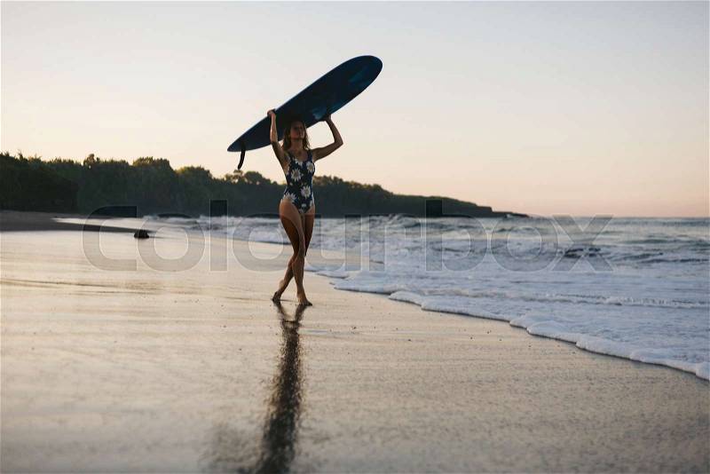 Sportswoman in swimming suit with blue surfing board walking on beach, stock photo