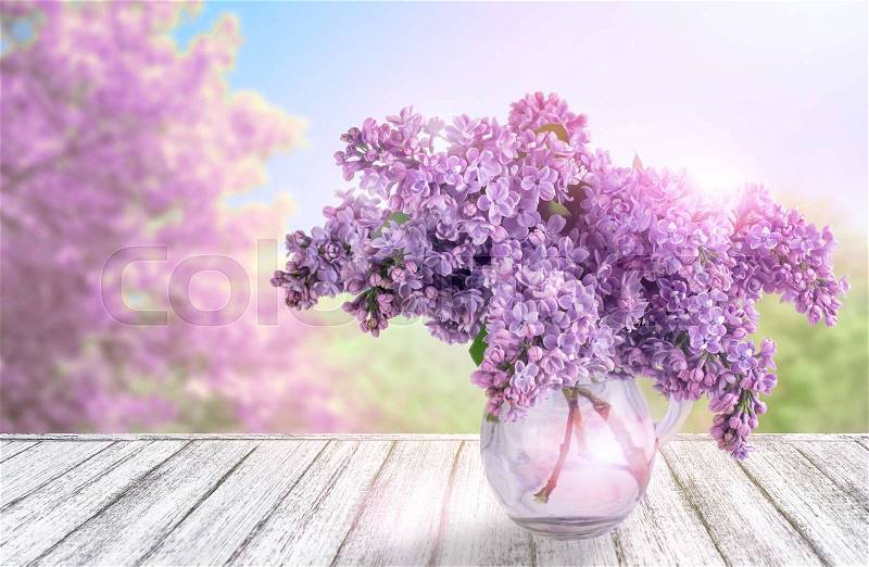 Wooden table in Shabby Chic style with lilac flowers in glass vase and spring trees against the blue sky, stock photo