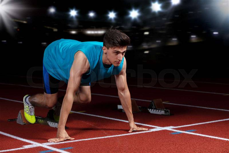 Sprinter leaving starting blocks on the running track in front of big modern stadium with lights and flares. Explosive start, stock photo