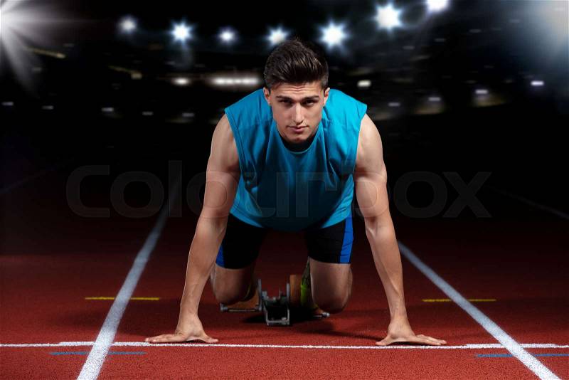 Sprinter leaving starting blocks on the running track in front of big modern stadium with lights and flares. Explosive start, stock photo