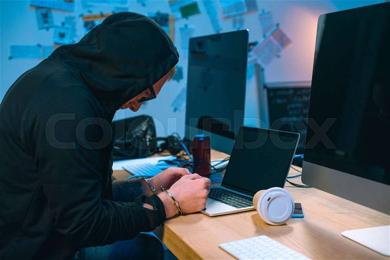 Handcuffed hacker in hoodie in front of laptop at workplace, stock photo