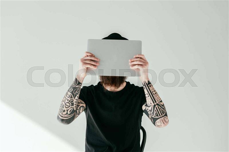 Bearded tattooed man covering face with laptop on white, stock photo