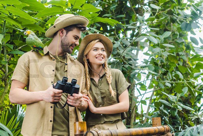 Happy young couple in safari suits with parrot in rainforest, stock photo