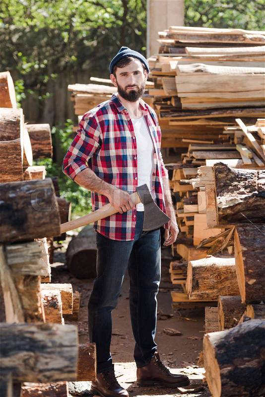 Lumberjack in checkered shirt standing with axe between logs at sawmill , stock photo