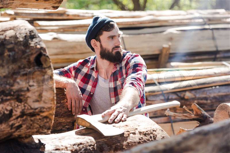 bearded lumberjack in checkered shirt with axe looking away at sawmill , stock photo