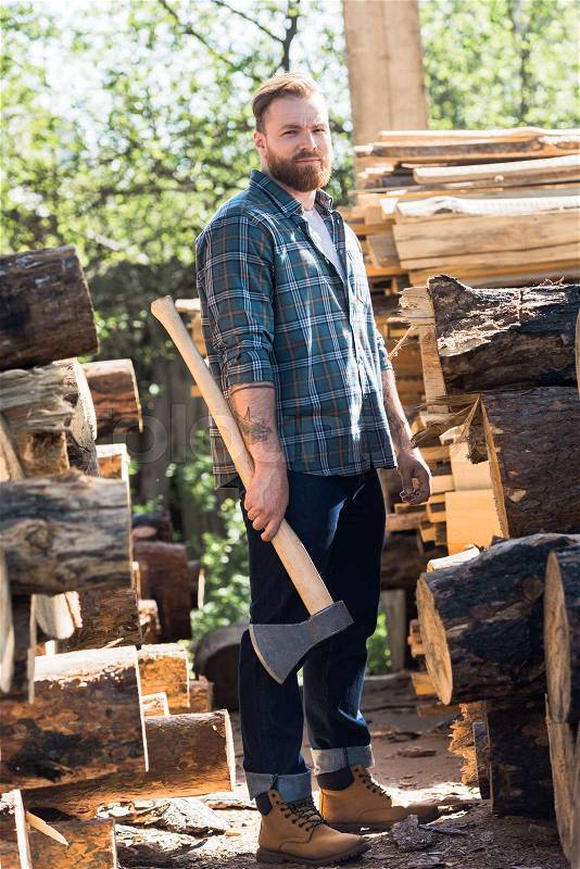 Bearded lumberjack in checkered shirt standing with axe at sawmill , stock photo