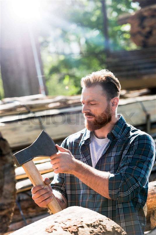 Bearded lumberjack in checkered shirt touching blade of axe at sawmill , stock photo