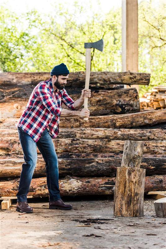 Bearded lumberjack in checkered shirt chopping log at sawmill , stock photo
