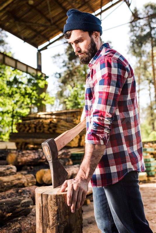 Bearded lumberjack in checkered shirt preparing to chop log at sawmill , stock photo