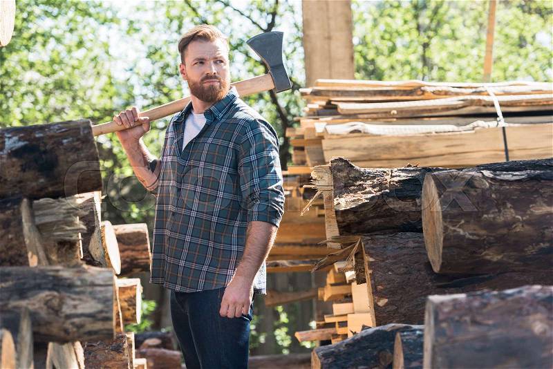 Lumberjack in checkered shirt holding axe on shoulder between logs at sawmill , stock photo