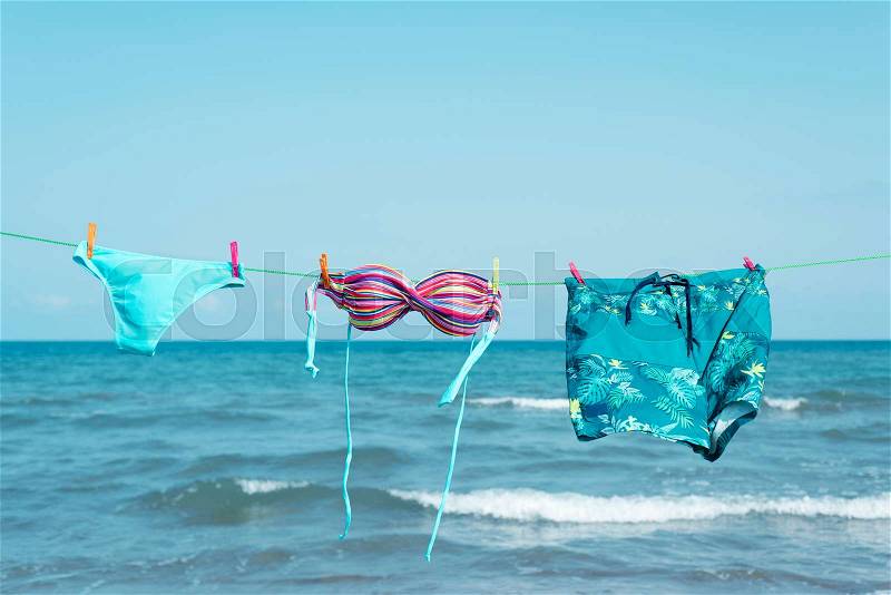 The two pieces of a colorful bikini and a pair of blue swimming trunks hanging on a clothes line on the beach, with the sea in the background, stock photo