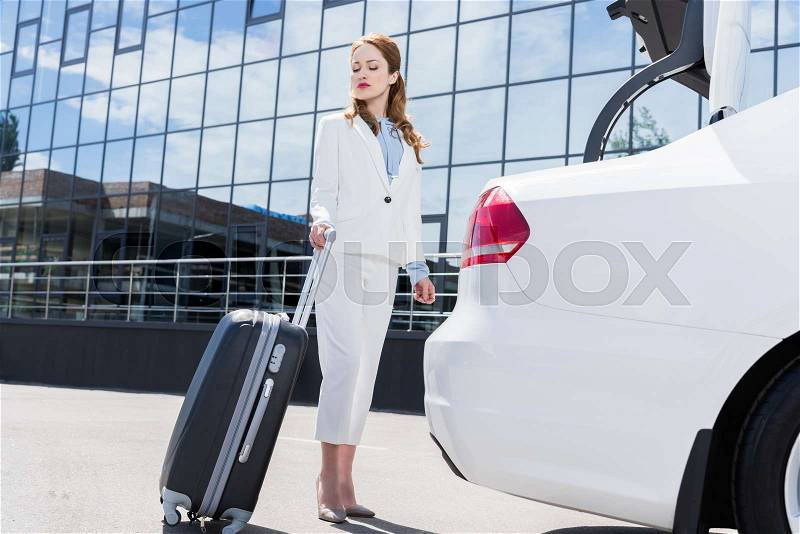 Side view of businesswoman in white suit with suitcase standing at car on street, stock photo