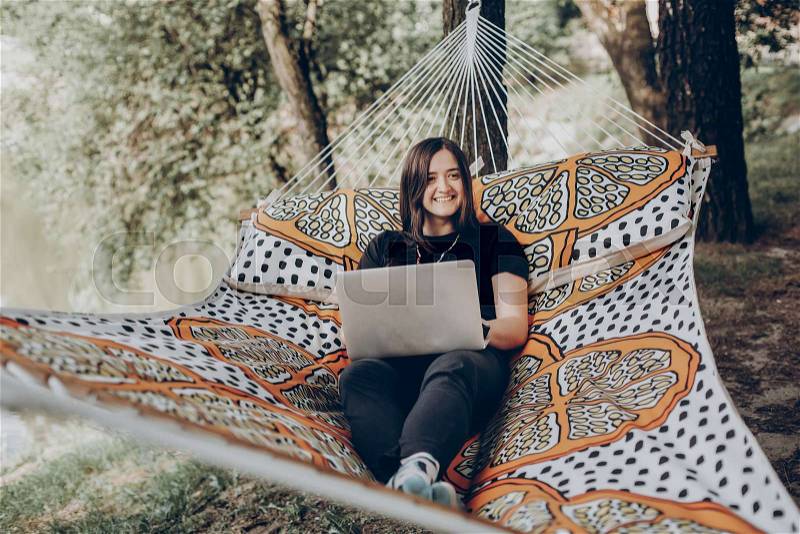 Beautiful brunette woman relaxing on hammock outdoors near lake in the forest, freelancer working in the park while resting in hammock, freelance concept, stock photo