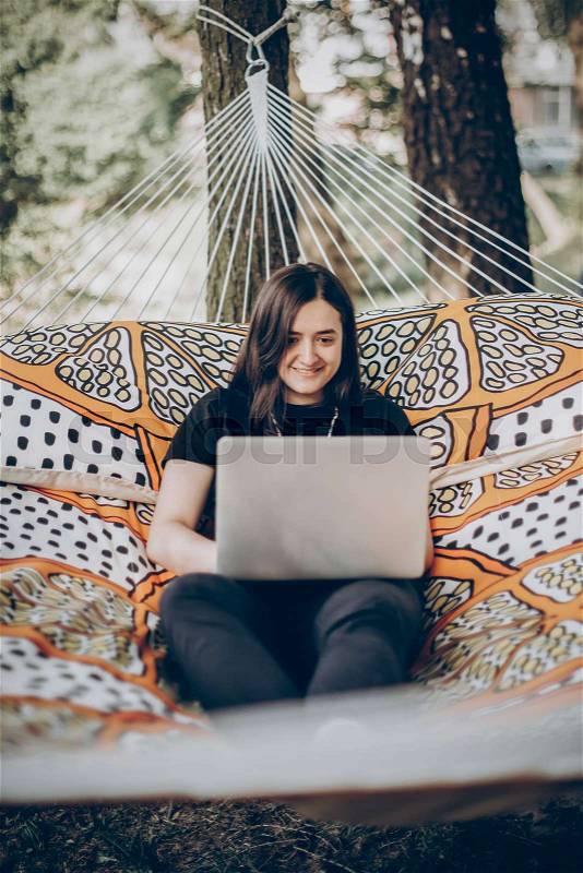 Beautiful brunette woman relaxing on hammock outdoors near lake in the forest, freelancer working in the park while resting in hammock, freelance concept, stock photo