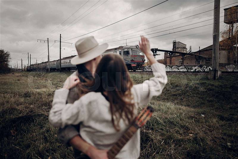 Sensual hipster couple, gypsy woman in boho clothes and musician with guitar wave at passing train, stock photo