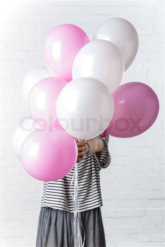 Woman holding pink and white balloons in front of her face on white brick wall background, stock photo