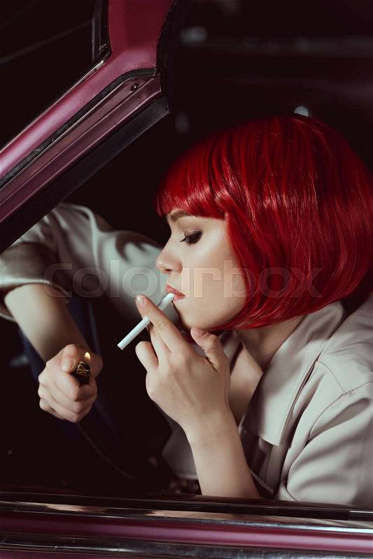 Side view of beautiful young woman in red wig smoking cigarette in car, stock photo