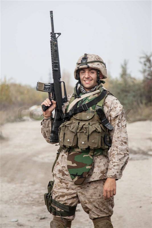 Smiling army soldier, United States Marine Corps infantry shooter in camo battle uniform, protected with body armor and helmet, posing with assault rifle in hands ..., stock photo
