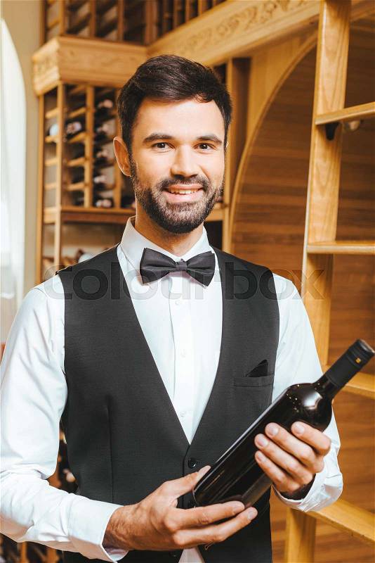 Smiling young wine steward with bottle of luxury wine at wine store, stock photo