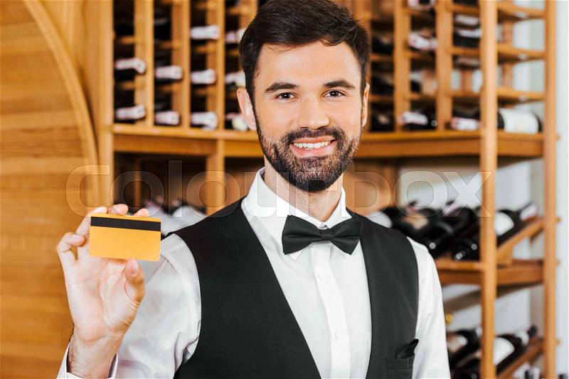 Smiling young wine steward holding golden card at wine store, stock photo
