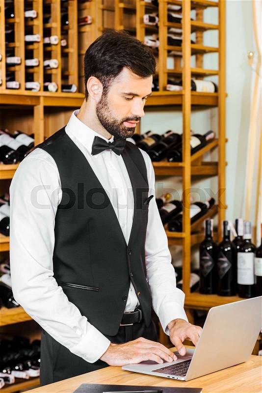 Handsome young wine steward working with laptop at wine store, stock photo