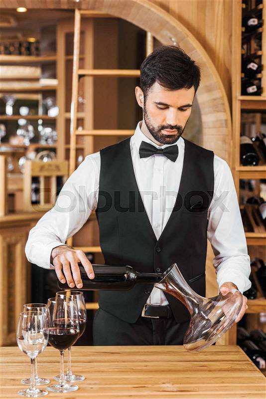 Handsome young wine steward pouring wine into decanter at wine store, stock photo