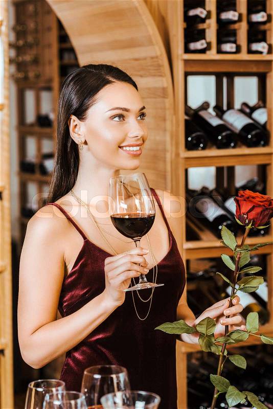 Beautiful woman in red dress with wine and rose looking at side at wine store, stock photo