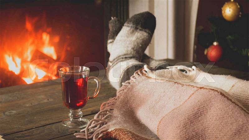 Toned image of woman in woolen socks lying under blanket and drinking tea at burning fireplace, stock photo