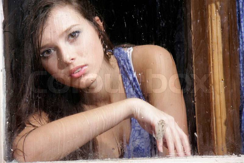 Young sad woman behind wet window, stock photo