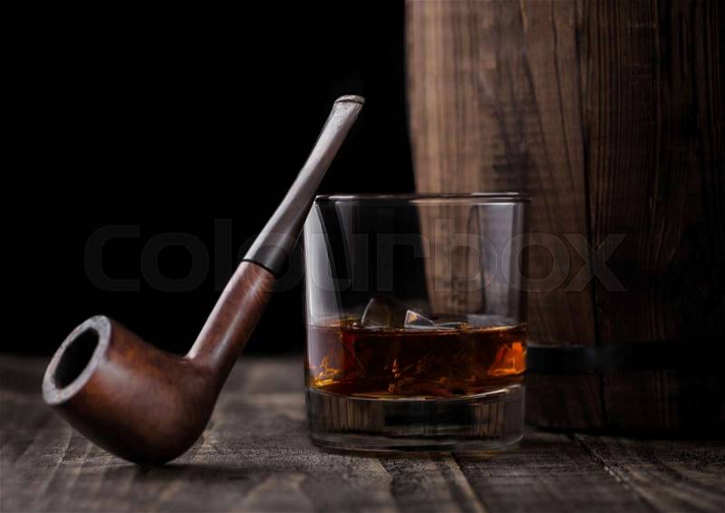 Glass of whiskey with ice cubes and vintage smoking pipe next to wooden barrel. Cognac and brandy drink, stock photo