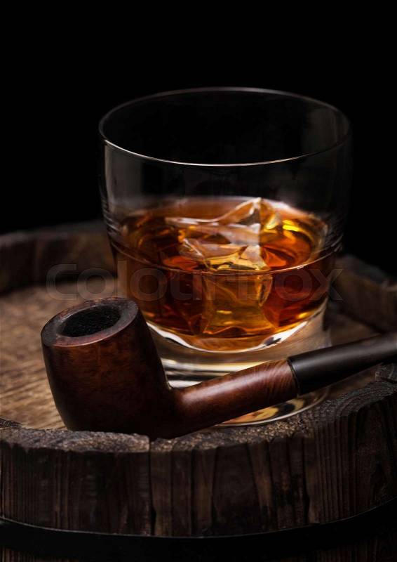 Glass of whiskey with ice cubes and vintage smoking pipe on top of wooden barrel. Cognac and brandy drink, stock photo