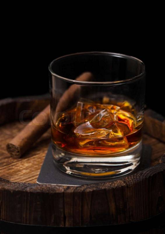 Glass of whiskey with ice cubes and cigar on top of wooden barrel. Cognac and brandy drink, stock photo
