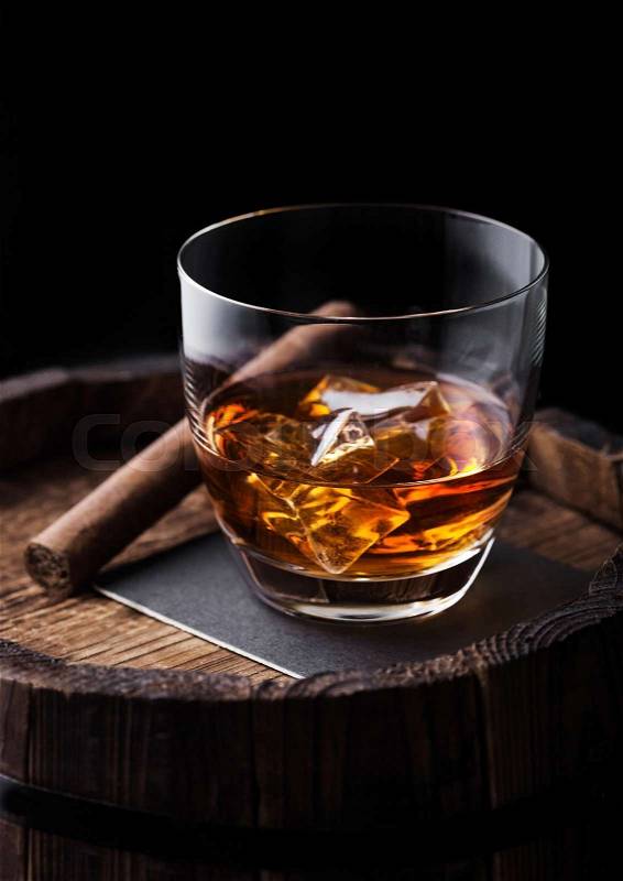 Glass of whiskey with ice cubes and cigar on top of wooden barrel. Cognac and brandy drink, stock photo