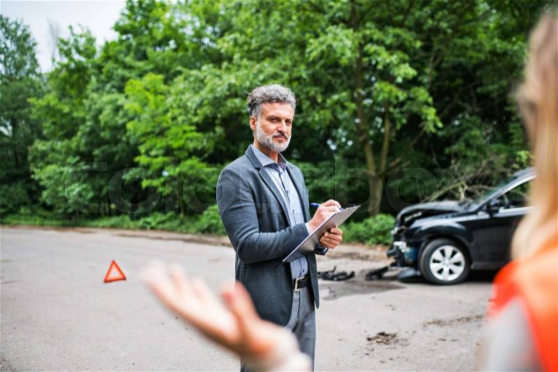 A man insurance agent talking to an unrecognizable woman outside on the road after a car accident, stock photo