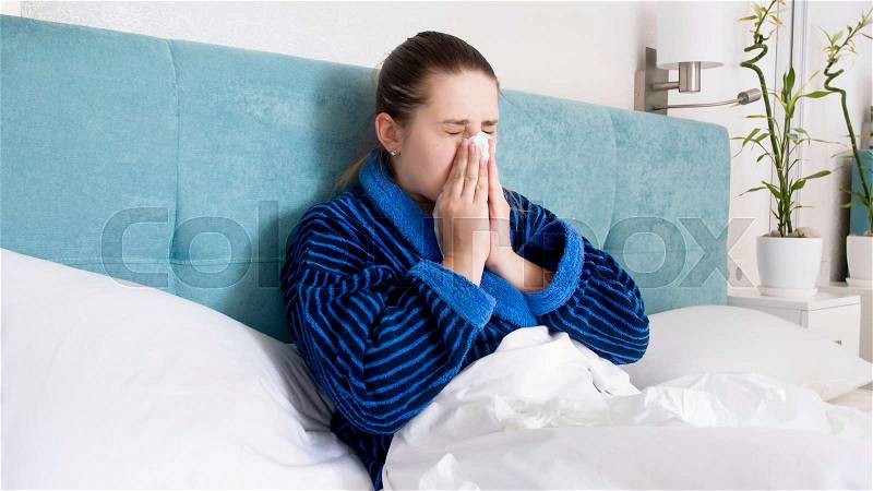 Portrait of young woman feeling unwell lying in bed and blowing nose in paper towel, stock photo