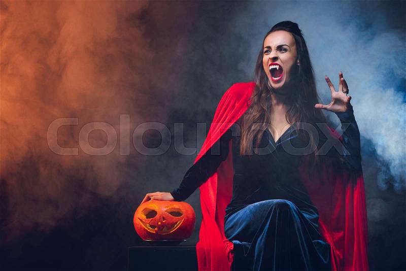 Mystic woman in vampire costume holding jack o lantern on darkness with smoke , stock photo