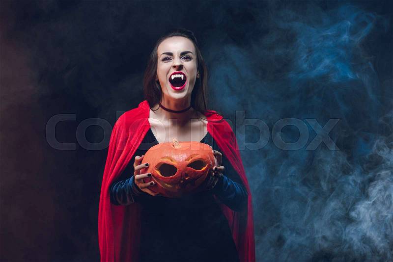 Mystic woman in vampire costume holding jack o lantern on dark background with smoke , stock photo