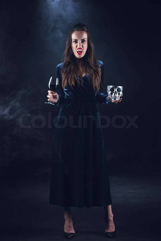 Vampire woman holding skull and wineglass with blood on dark background with smoke , stock photo