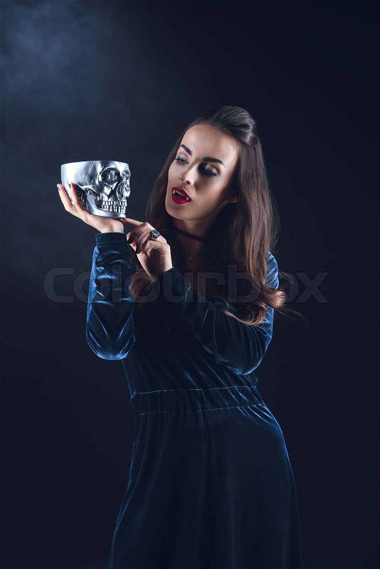 Vampire woman holding metal skull on dark background with smoke , stock photo