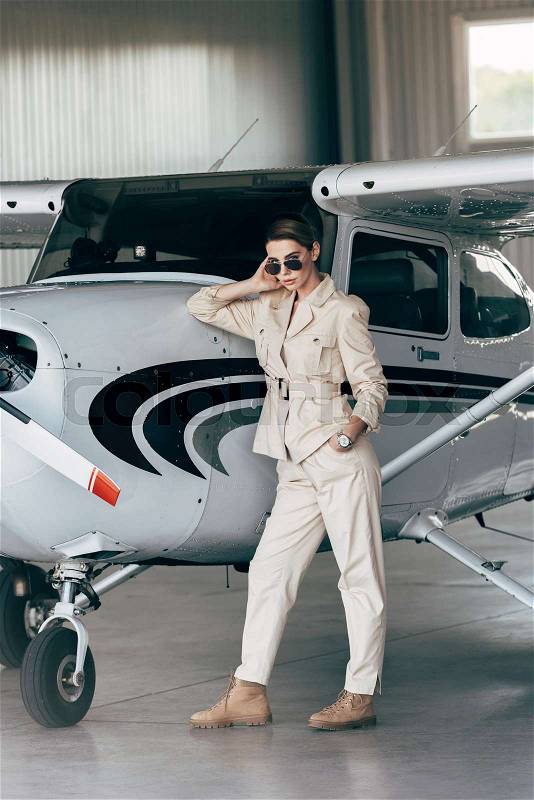 Fashionable young woman in sunglasses and jacket posing near aircraft in hangar, stock photo