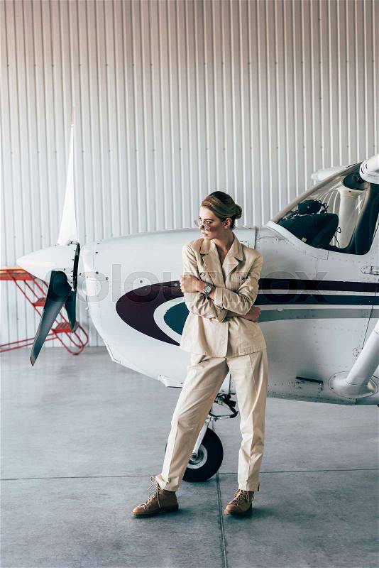 Fashionable woman in sunglasses and jacket posing near aircraft in hangar, stock photo
