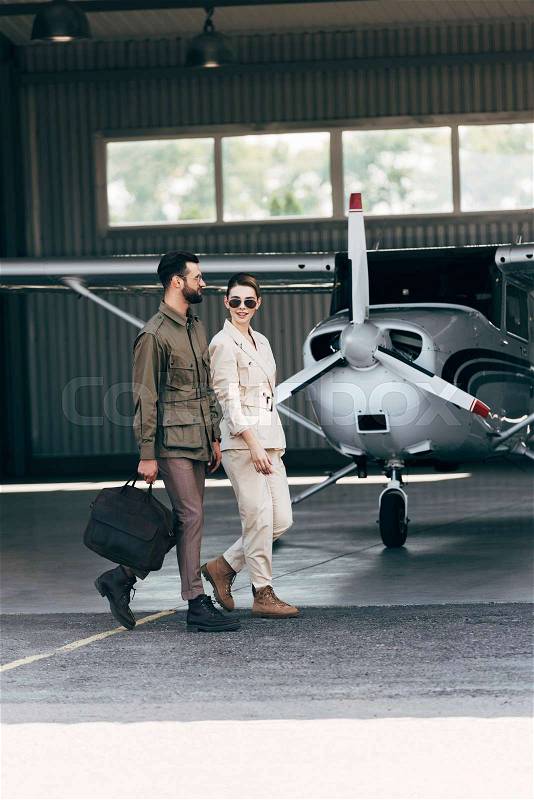 Stylish man carrying bag and walking with girlfriend near hangar with plane , stock photo